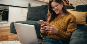 A woman texting on her phone with her laptop on her lap and back against the sofa