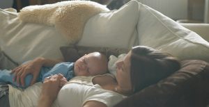 A mother laying asleep on the sofa with her baby sleeping on top of her