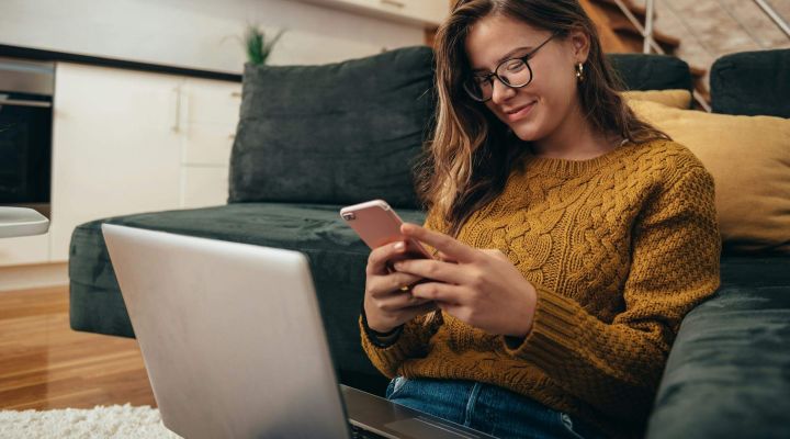 A woman texting on her phone with her laptop on her lap and back against the sofa