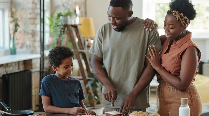 Young man cutting bread for sandwiches by table in the kitchen for his affectionate wife and their adorable little son surrounding him