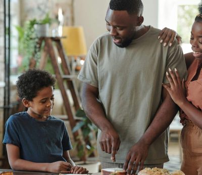Young man cutting bread for sandwiches by table in the kitchen for his affectionate wife and their adorable little son surrounding him