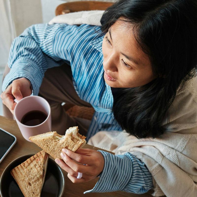 A woman drinking a coffee and eating a sandwich while wrapped up in a blanket